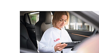 Zu sehen ist eine Pflegefachkraft in einem Auto der Sozialstation, den Blick auf ein Tablet gerichtet. 
